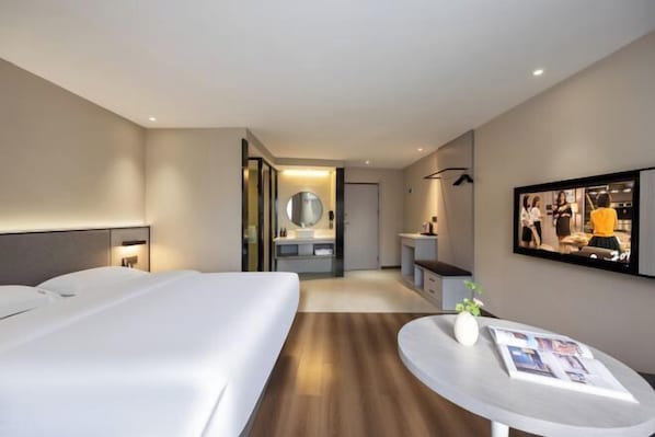 Une chambre d’hôtel comprenant un lit, une table de chevet, une table ronde avec un vase et un magazine, un miroir et un téléviseur fixé au mur.