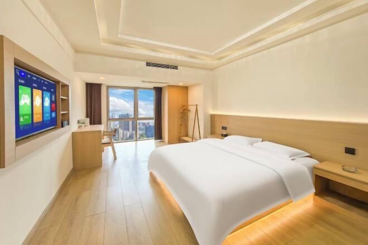 A modern hotel room with a large bed, a TV mounted on the wall, and a view of the city through the window.