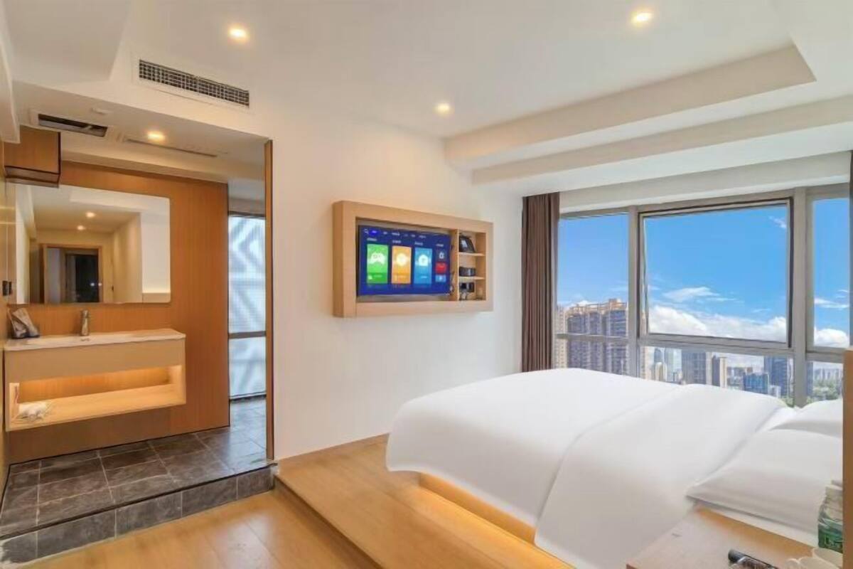 A modern hotel room with a large bed, a mounted TV, a bathroom with a vanity, and a city view.