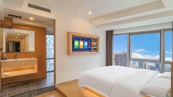 A modern hotel room with a large bed, a mounted TV, a bathroom with a vanity, and a city view.