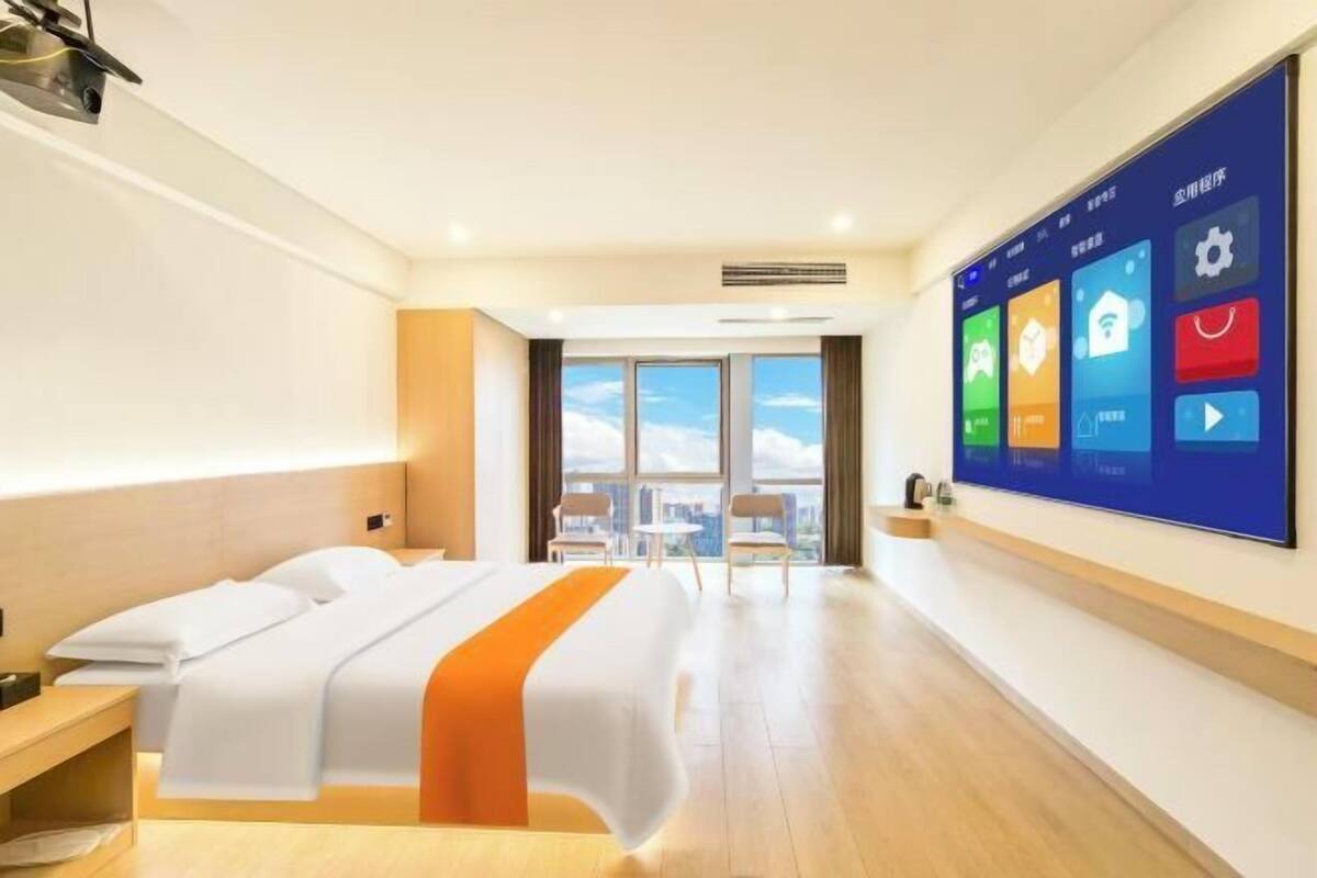 A hotel room with a bed, a large digital screen displaying room controls, a view of the city through the window, and a wooden floor.
