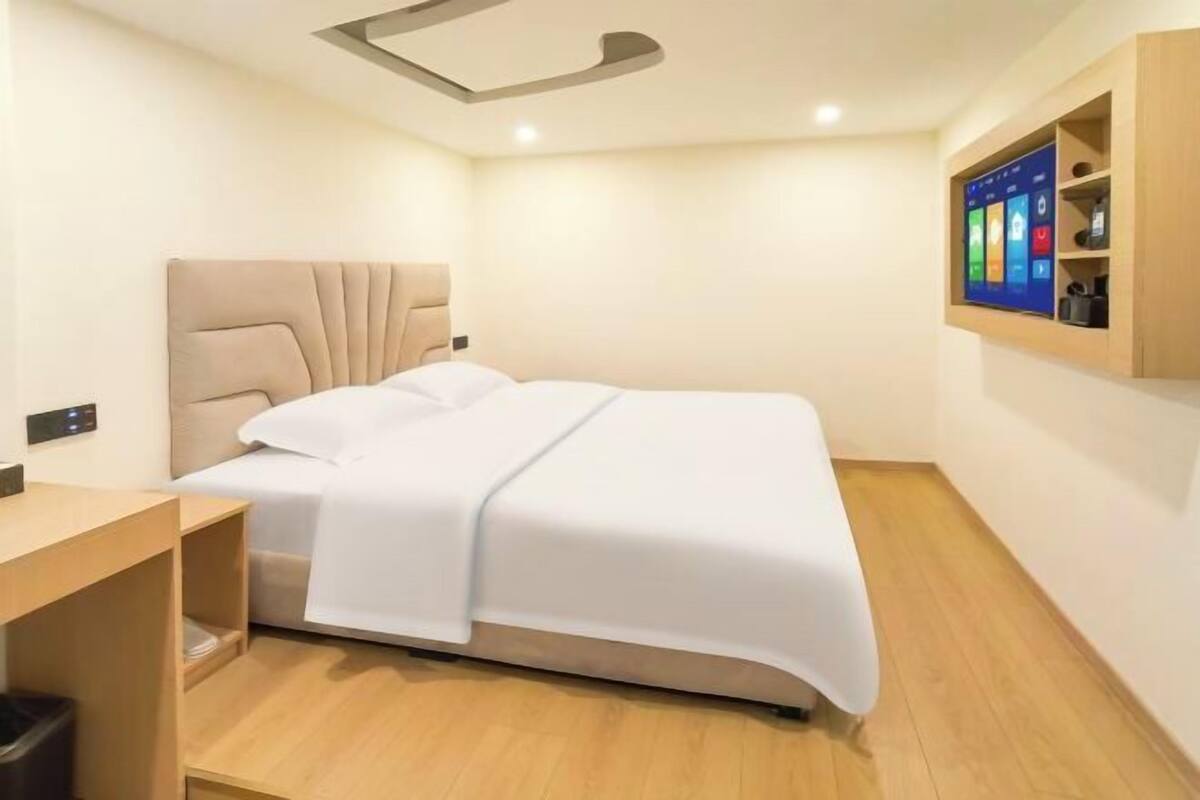 A modern hotel room with a bed, a wooden desk, a built-in shelf with a TV, and a ceiling light fixture.