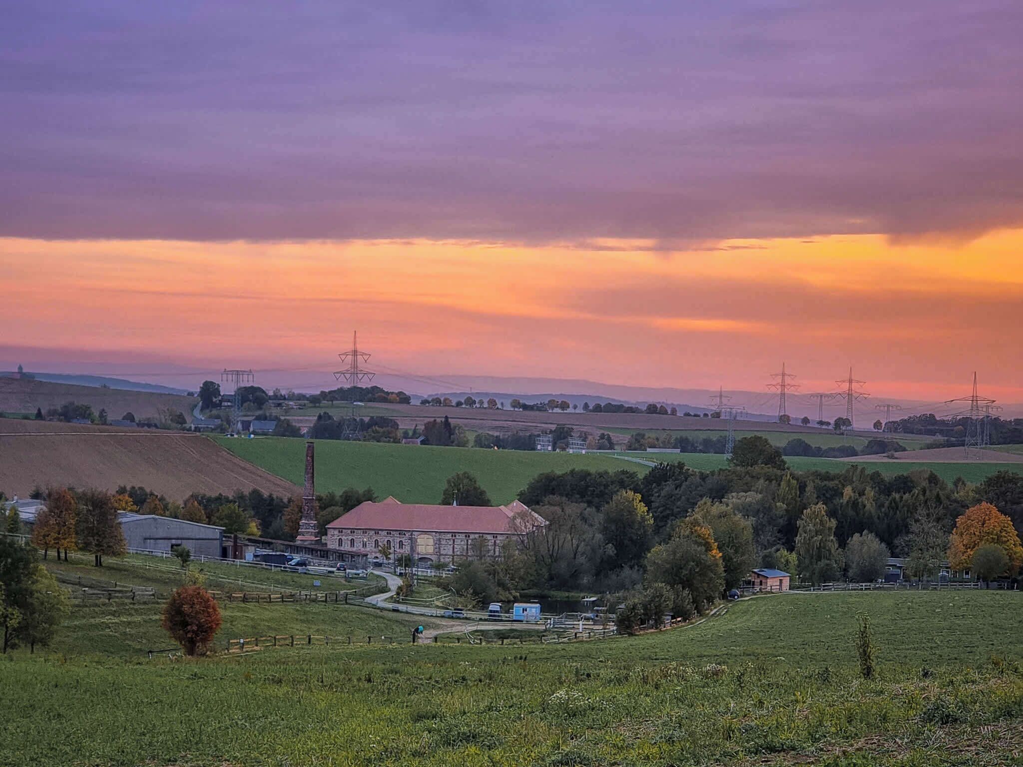 Un paysage rural caractérisé par un bâtiment imposant, de vastes champs ouverts et des lignes électriques qui disparaissent à l’horizon.