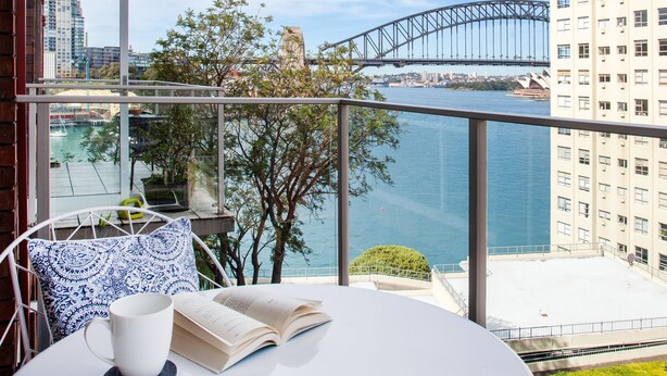 A balcony with a view of the Sydney Harbour Bridge, a table with a book and a mug, and a chair with a patterned cushion. - Harbour the Sun, McMahons Point Isyd (McMahons Point)