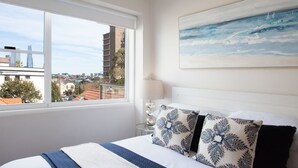 A hotel room with a large window offering a city view, a bed with patterned pillows, a bedside table with a lamp, and a painting on the wall. - Harbour the Sun, McMahons Point Isyd (McMahons Point)