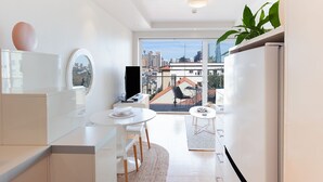 A modern kitchen with white cabinetry, a round mirror, and a dining area with a table and chairs. A large window offers a view of the city skyline. - Harbour the Sun, McMahons Point Isyd (McMahons Point)