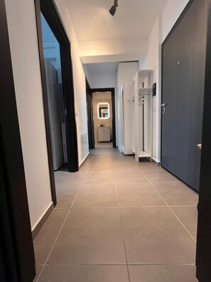 Un couloir avec un sol carrelé, une porte sur la droite et un miroir avec une lampe fixée au mur.