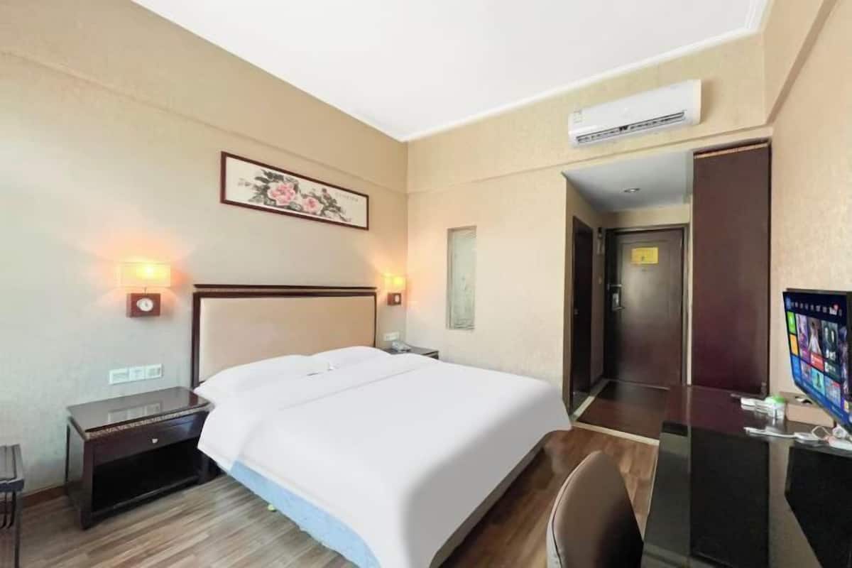 A hotel room with a bed, bedside tables, a desk with a computer, and an air conditioning unit.