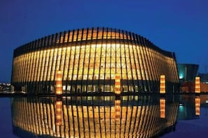 Um edifício grande e circular com fachada refletora, possivelmente um estádio ou arena, com reflexo nítido na água em frente.
