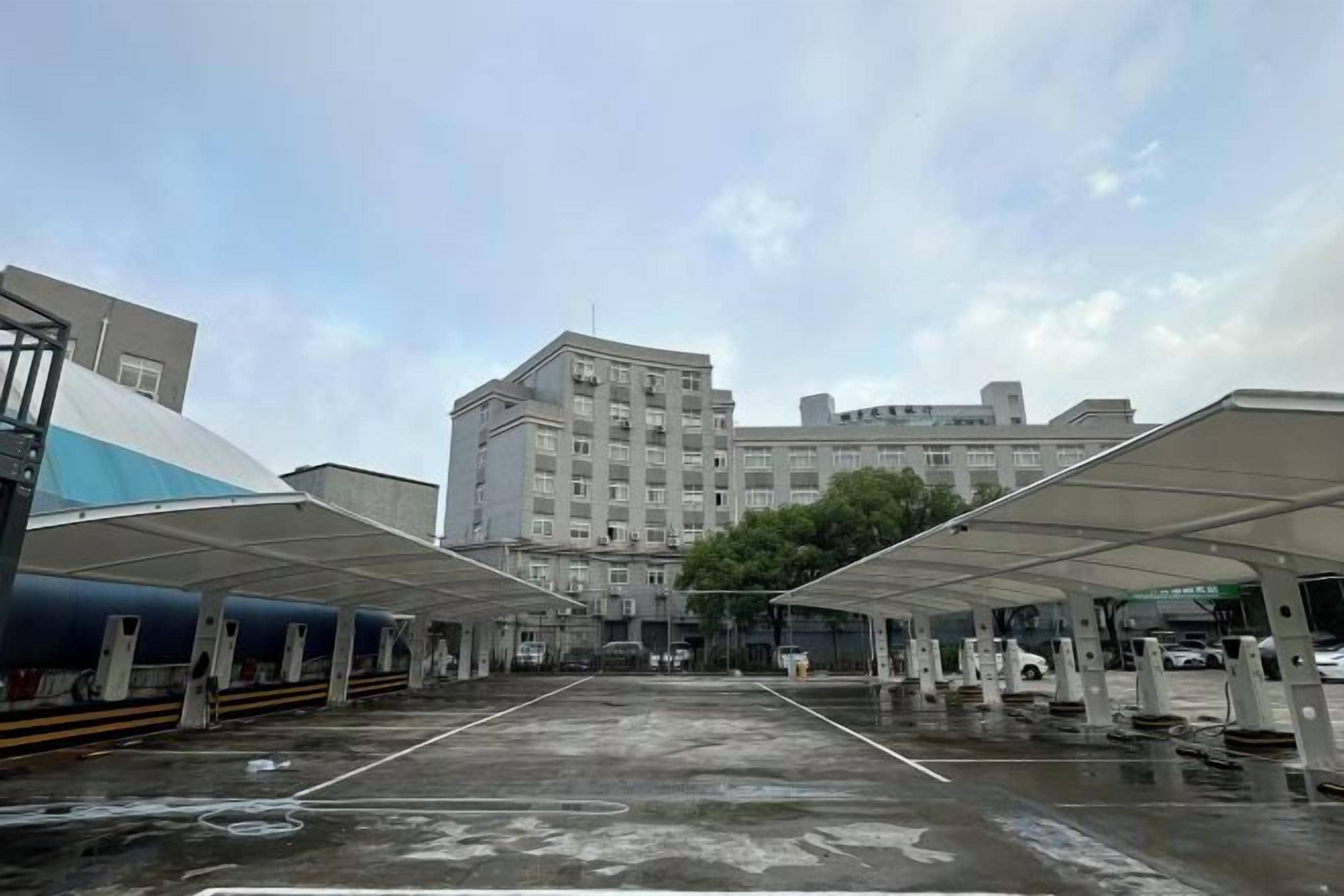 A parking area with covered canopies, a large building in the background, and a wet ground surface.