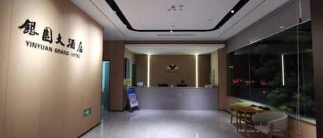 A hotel reception area with a reception desk, a seating area, and a large window providing a view of the outside.