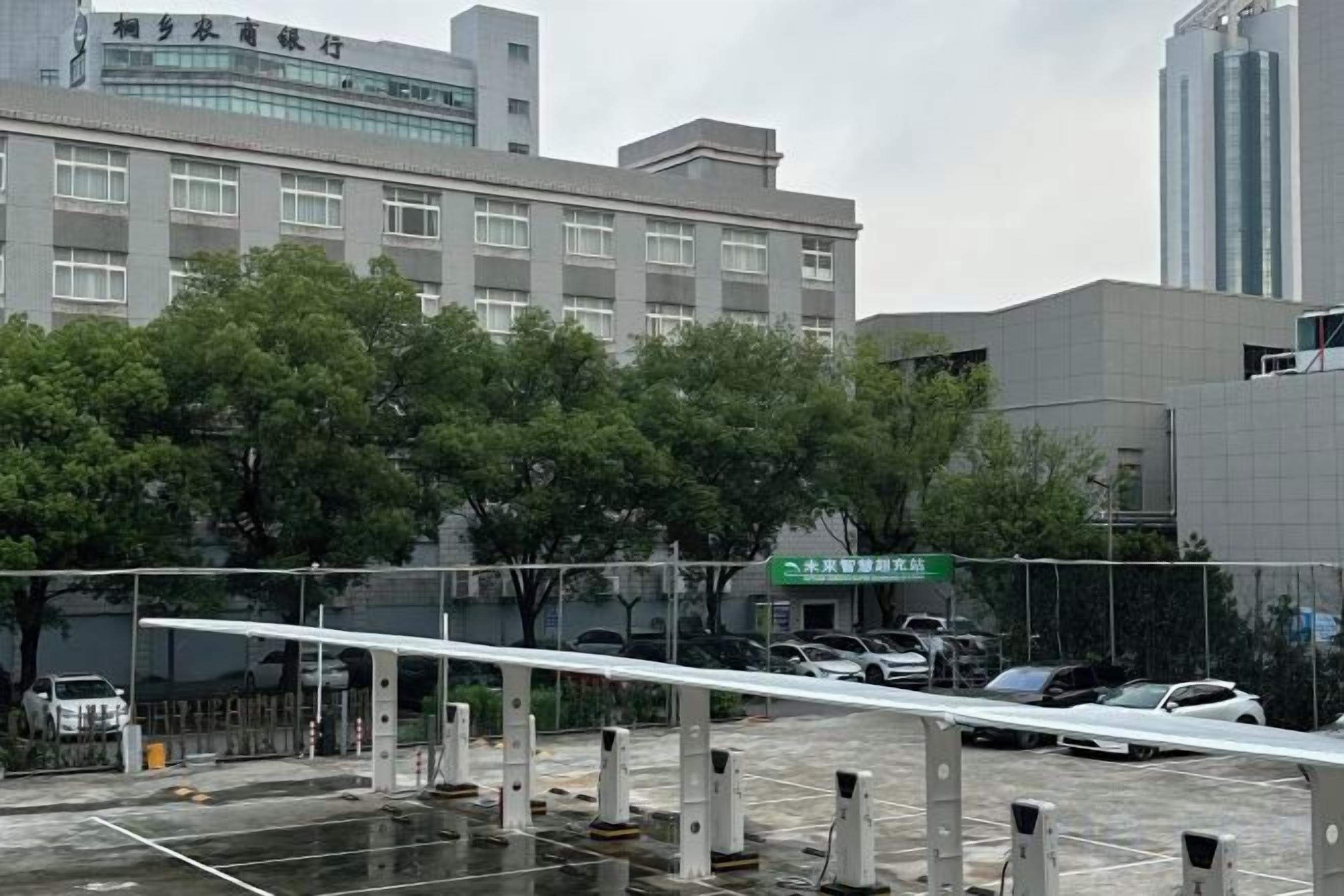 Un estacionamiento con una barrera blanca, árboles y un edificio de varios pisos al fondo.