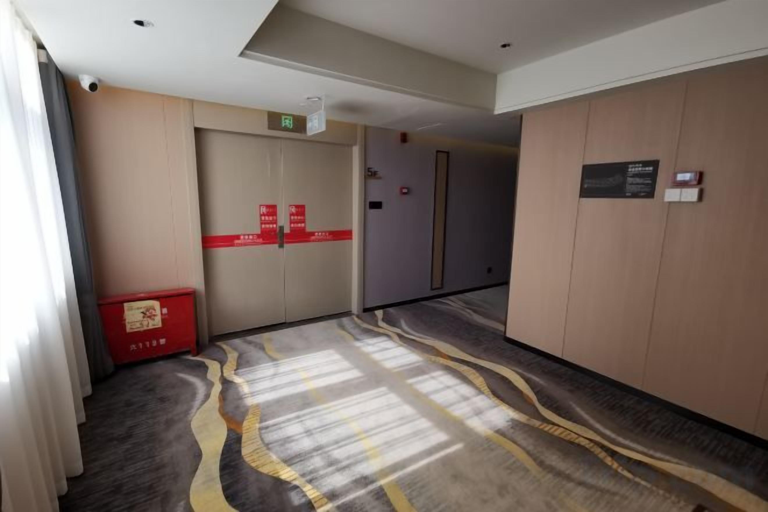 Un pasillo de hotel con piso alfombrado, una caja roja para almacenamiento y una puerta con un cartel.