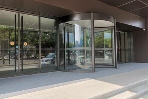 Una entrada moderna de edificio con puerta giratoria, puertas de vidrio y un área pavimentada que conduce a la entrada.