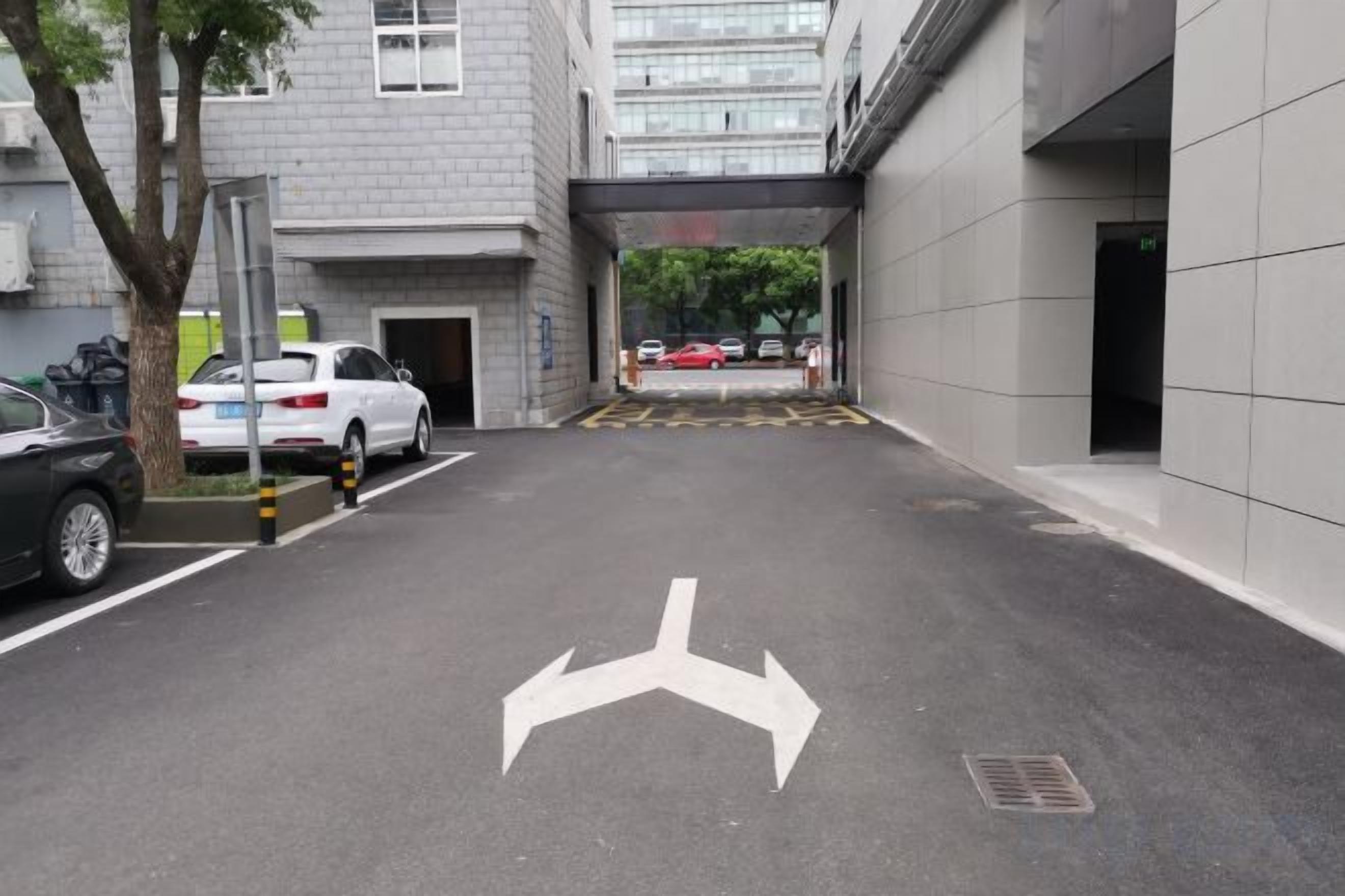 Un área de estacionamiento con flechas direccionales en el suelo, un árbol y la entrada a un edificio.