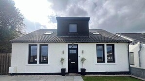 A single-story house with a black roof, white exterior walls, and a front door. There are two potted plants on either side of the door, and a small window above the door. - St Annes - Cosy Cottage in fife (Kirkcaldy)