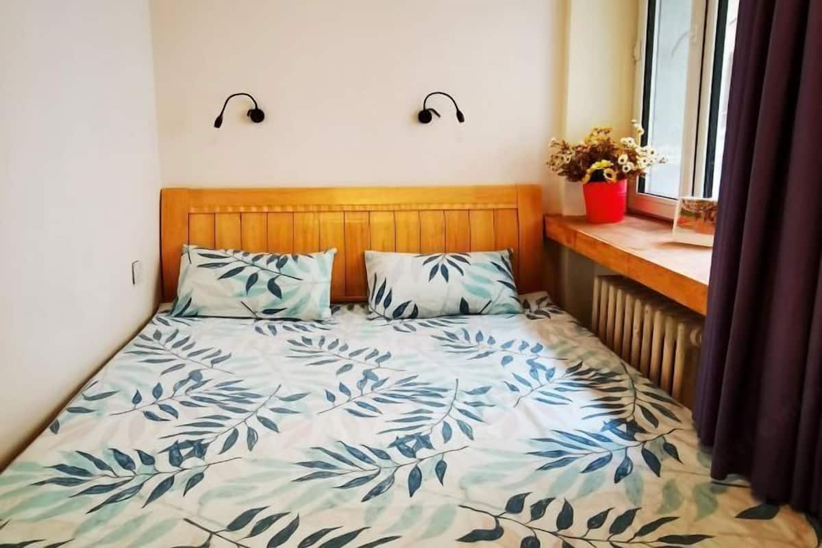 Une chambre à coucher avec une tête de lit en bois, deux haut-parleurs fixés au mur, une fenêtre avec vue et une plante en pot posée sur le rebord de la fenêtre.