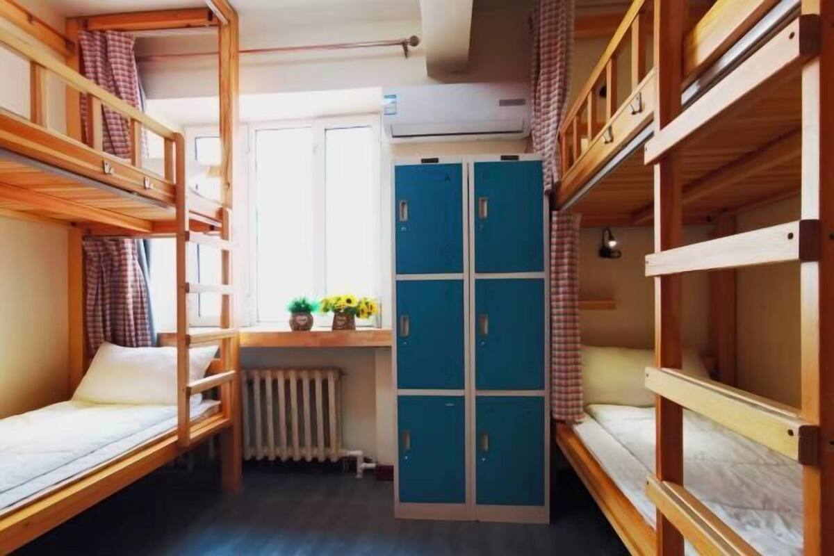 Une chambre de dortoir avec des lits superposés, un radiateur, une fenêtre avec des rideaux et une armoire bleue.