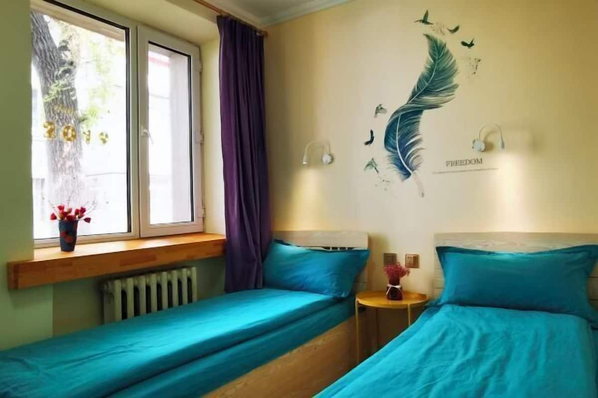 Une chambre avec deux lits simples, une fenêtre avec des rideaux, une fresque murale, une petite table avec un vase et un radiateur.