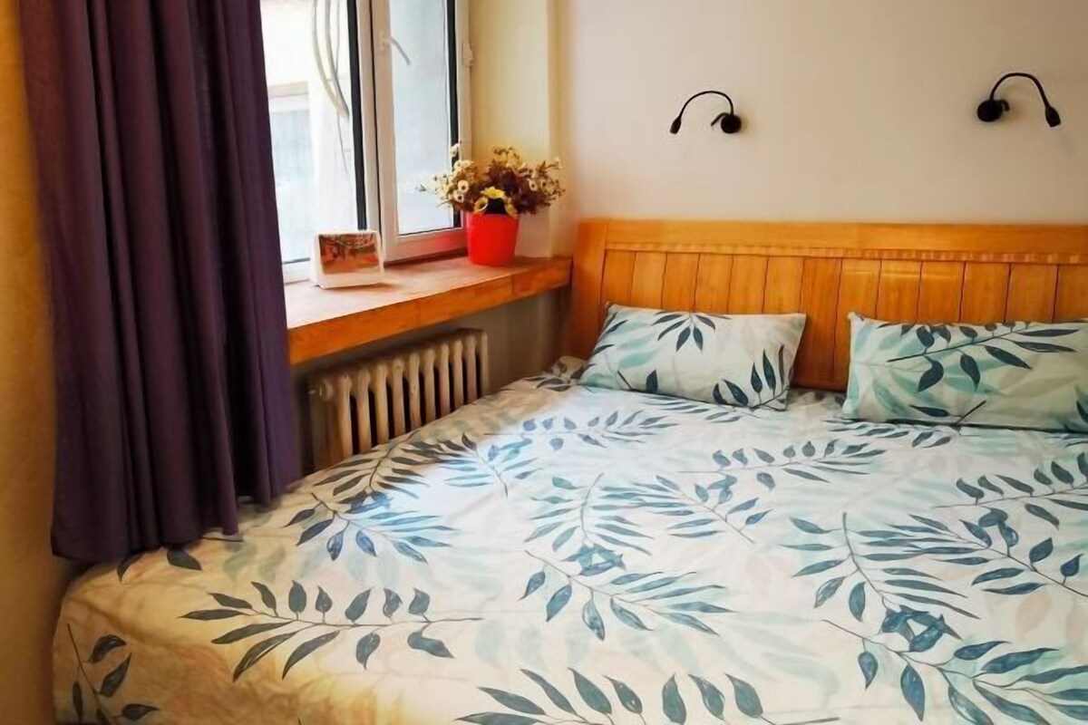 A bedroom with a bed, a window with curtains, a radiator, a wooden headboard, and a wall-mounted light fixture.
