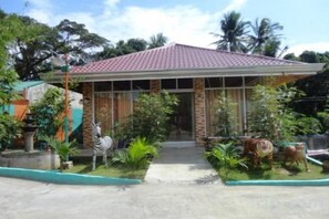 Uma casa com telhado de telhas vermelhas, varanda frontal e jardim com diversas plantas e uma estátua de zebra.
