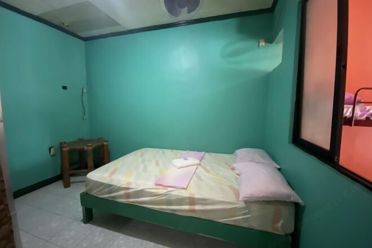 A small, single-bed room with teal walls, a wooden stool, and a window with a metal frame.