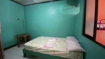Una habitación pequeña con cama individual, paredes verde azulado, un taburete de madera y una ventana con marco de metal.