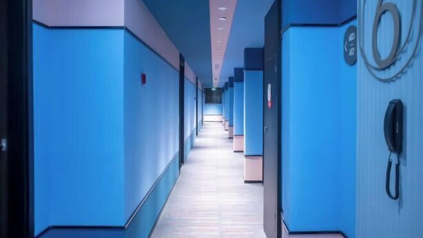 A long, narrow hallway with blue walls and a patterned floor, leading to a door with a phone on the right.