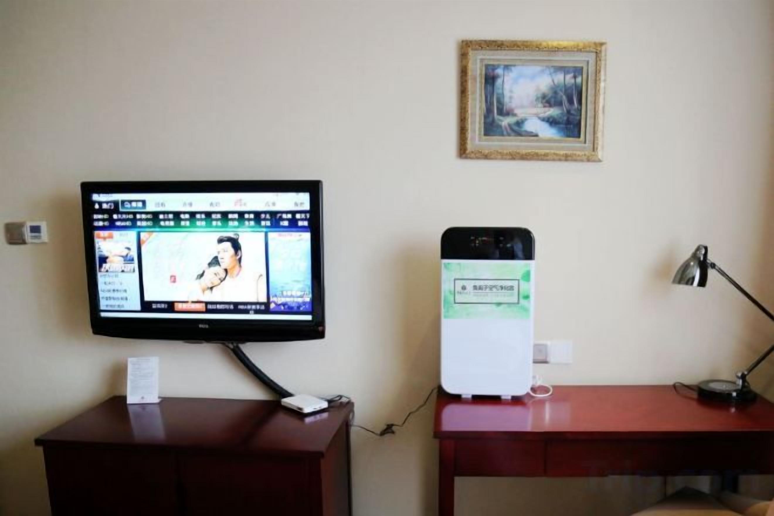 Ett rum med en tv på ett stativ, ett skrivbord med en lampa och en inramad tavla på väggen.