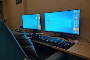 Two computer monitors on a desk with a keyboard and mouse, a chair, and a wall-mounted shelf. - Halcyon Hotel & Resort (Yingtan)