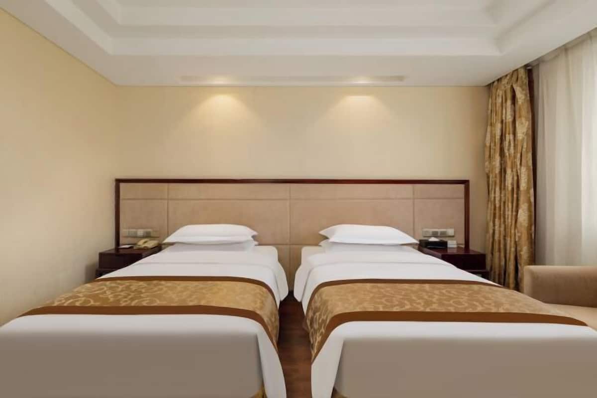 A hotel room with two single beds, a wooden headboard, bedside tables with lamps, and a beige carpet.
