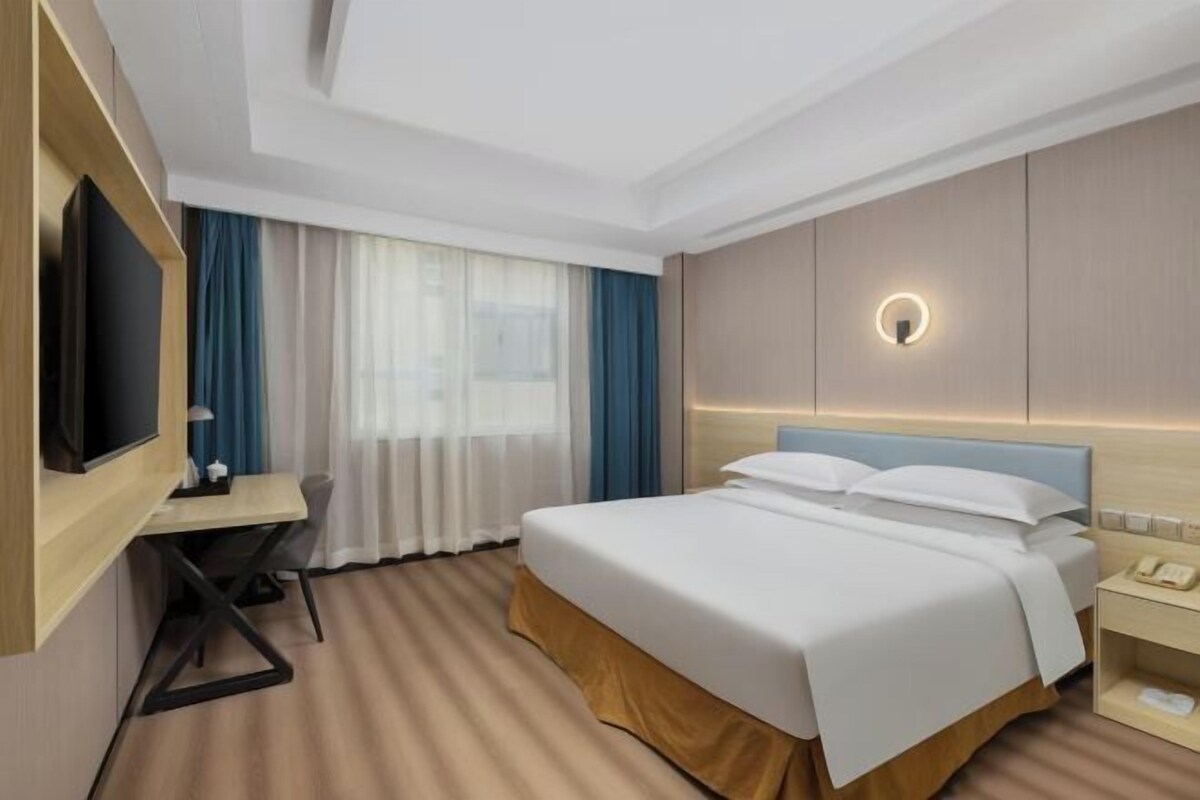 A hotel room with a bed, a desk, a TV, a bedside table, and a window with curtains.