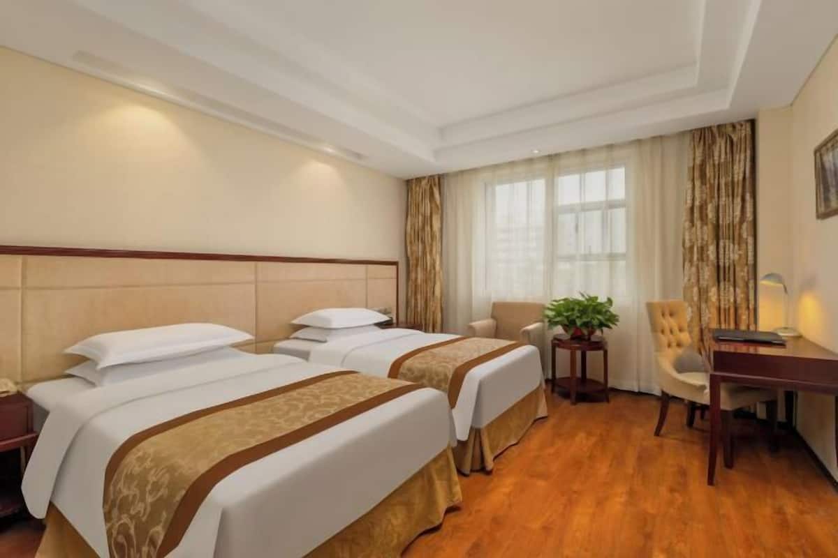 A hotel room with two beds, a desk, a chair, and a plant.