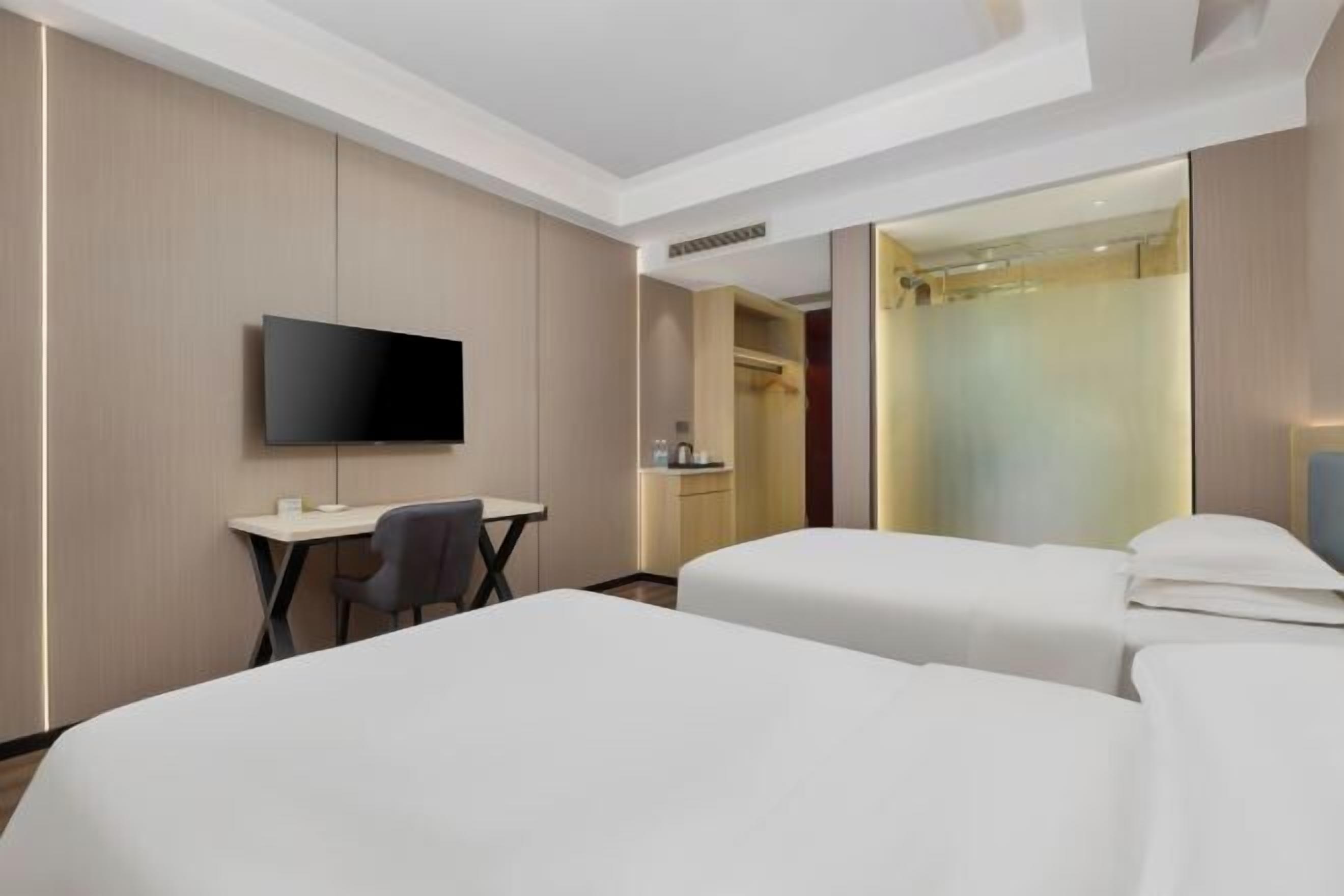A hotel room with two beds, a desk, a chair, a TV, and a shower area.