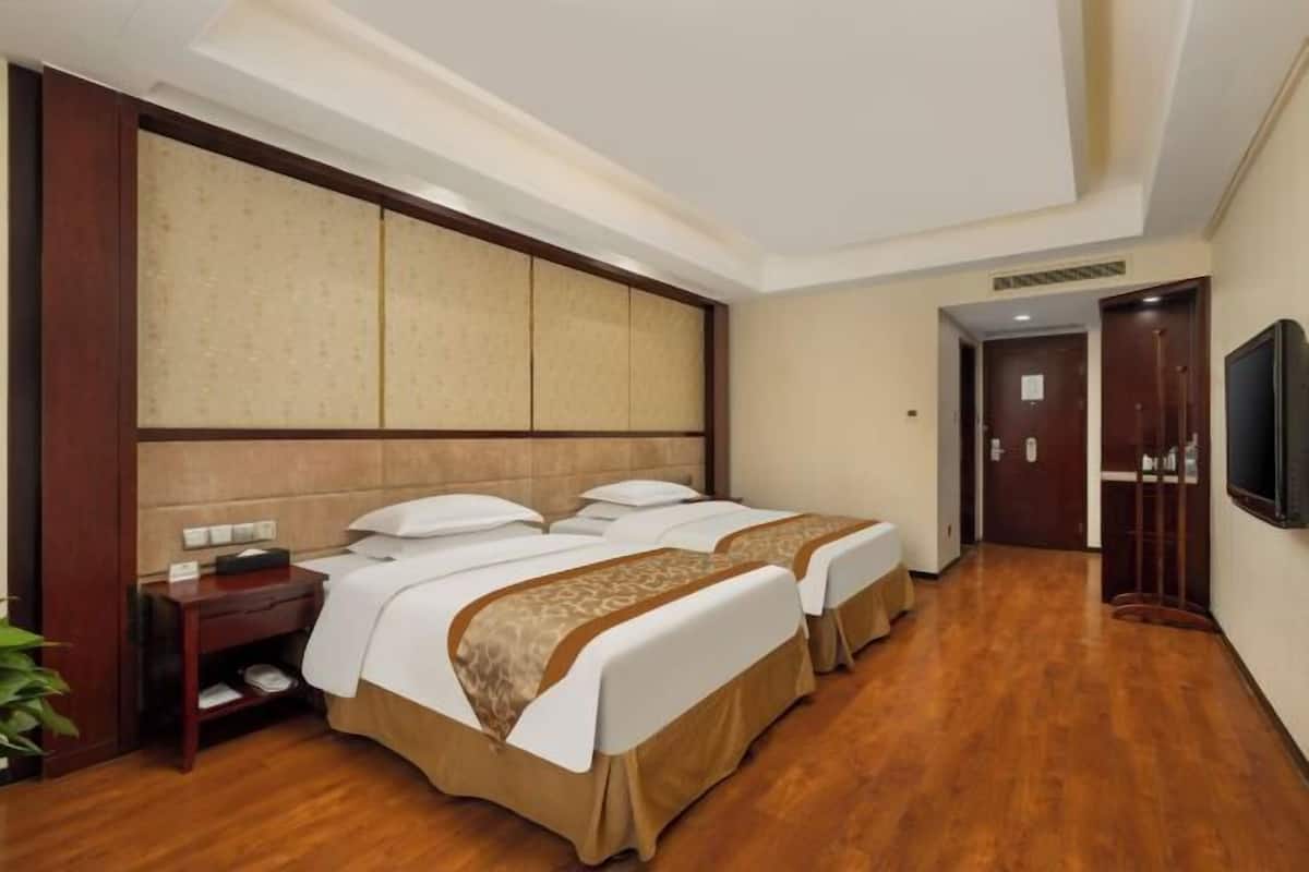 A hotel room with two beds, wooden flooring, a large wall panel, a TV mounted on the wall, and a door leading to another room.