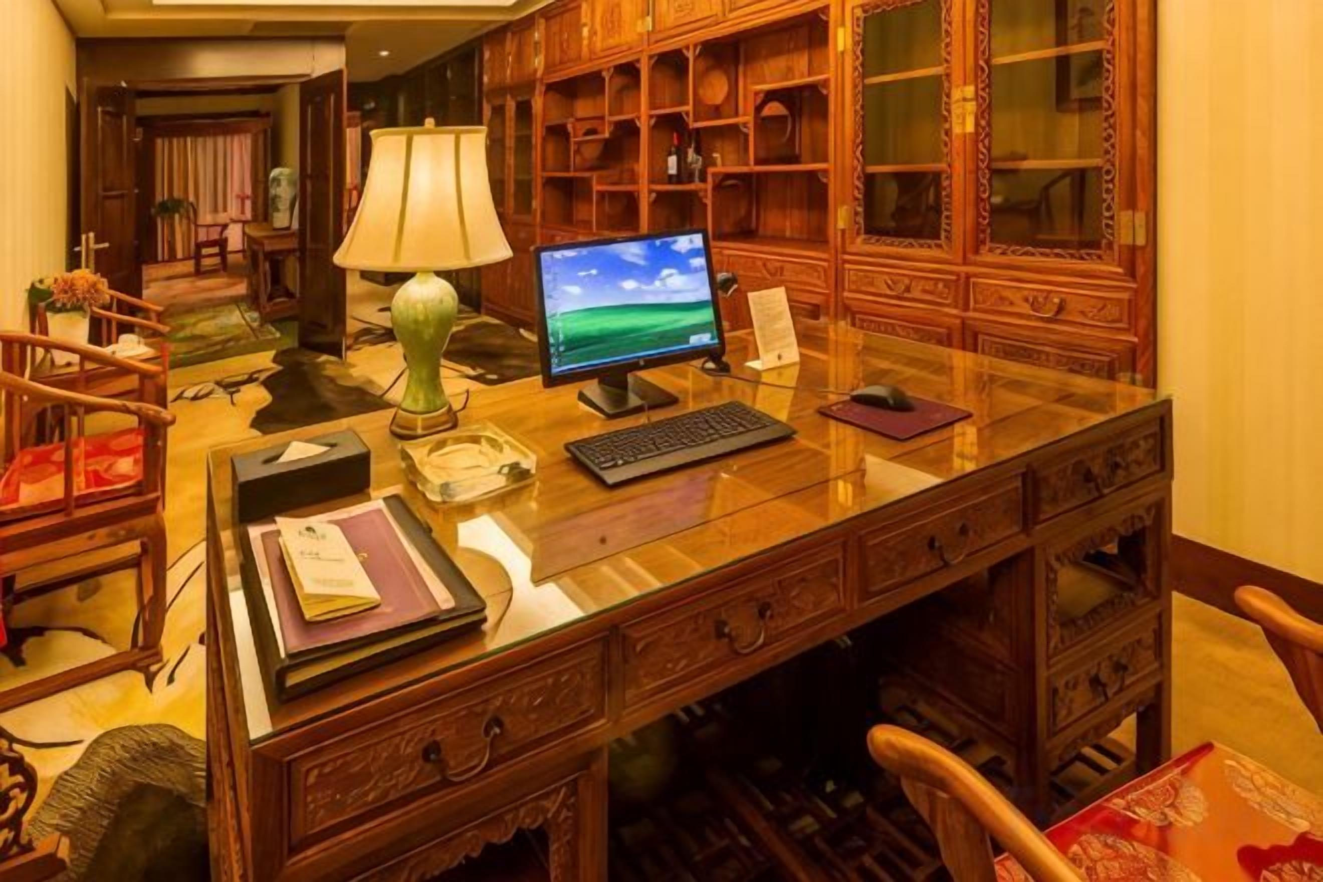 Un bureau en bois avec un ordinateur, une lampe et des livres, dans une pièce où se trouvent une bibliothèque et une chaise.