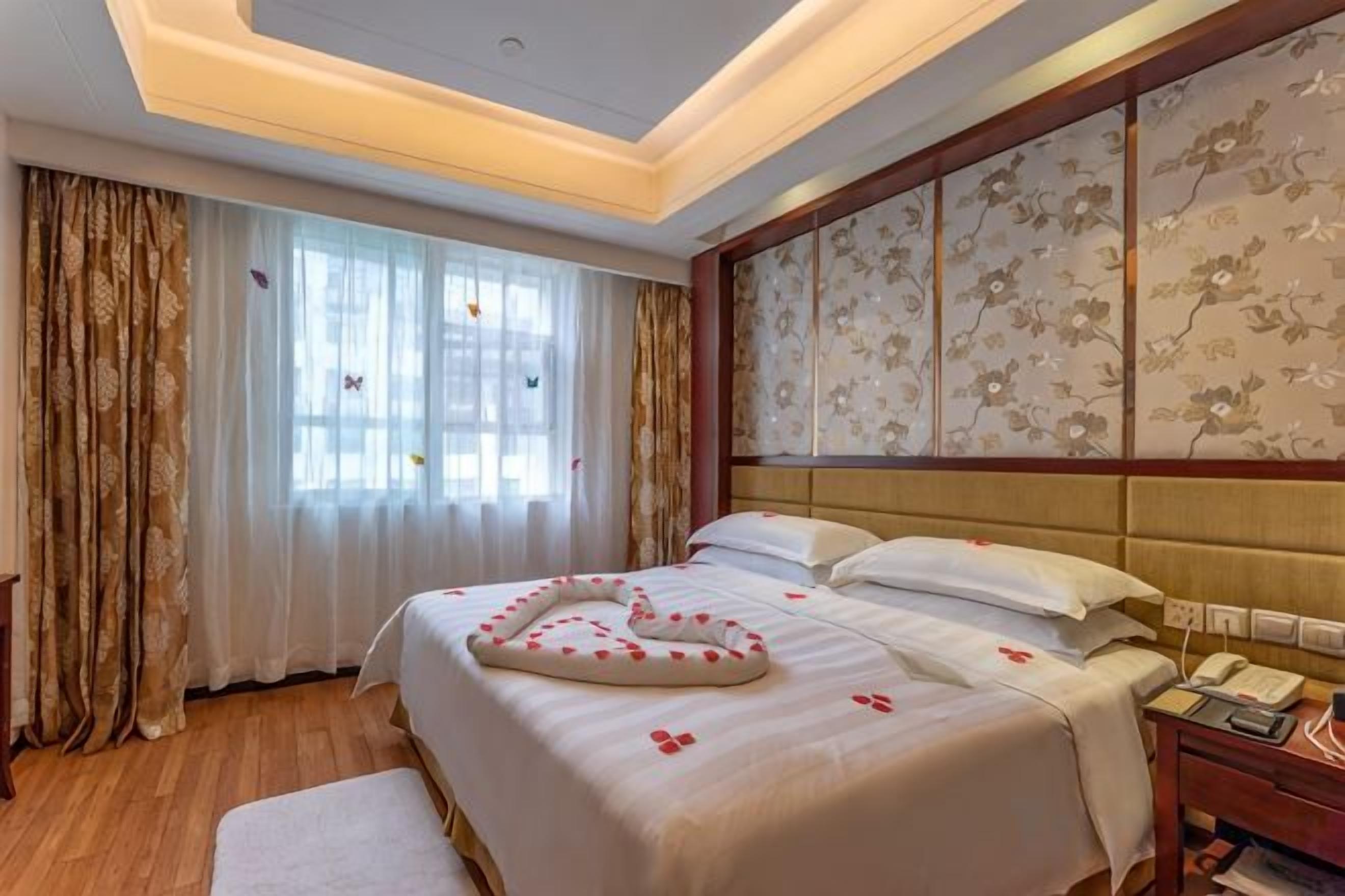 A hotel room with a large bed, a bedside table with a telephone, and a window with curtains.