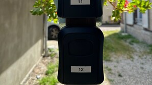 A parking meter with numbers 11 and 12, located in a narrow street with red flowers and a building in the background.
