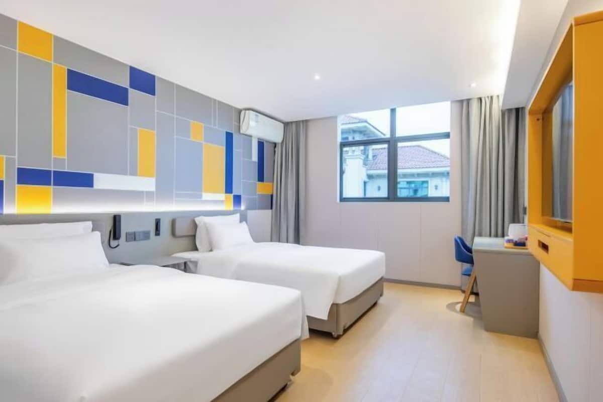 A hotel room with two beds, a desk, a chair, and a large window with a view of buildings.