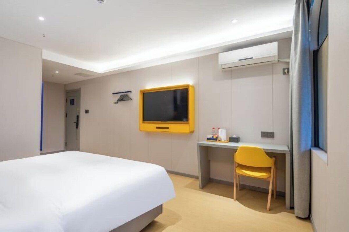 A hotel room with a bed, a desk, a chair, a television, and an air conditioning unit.