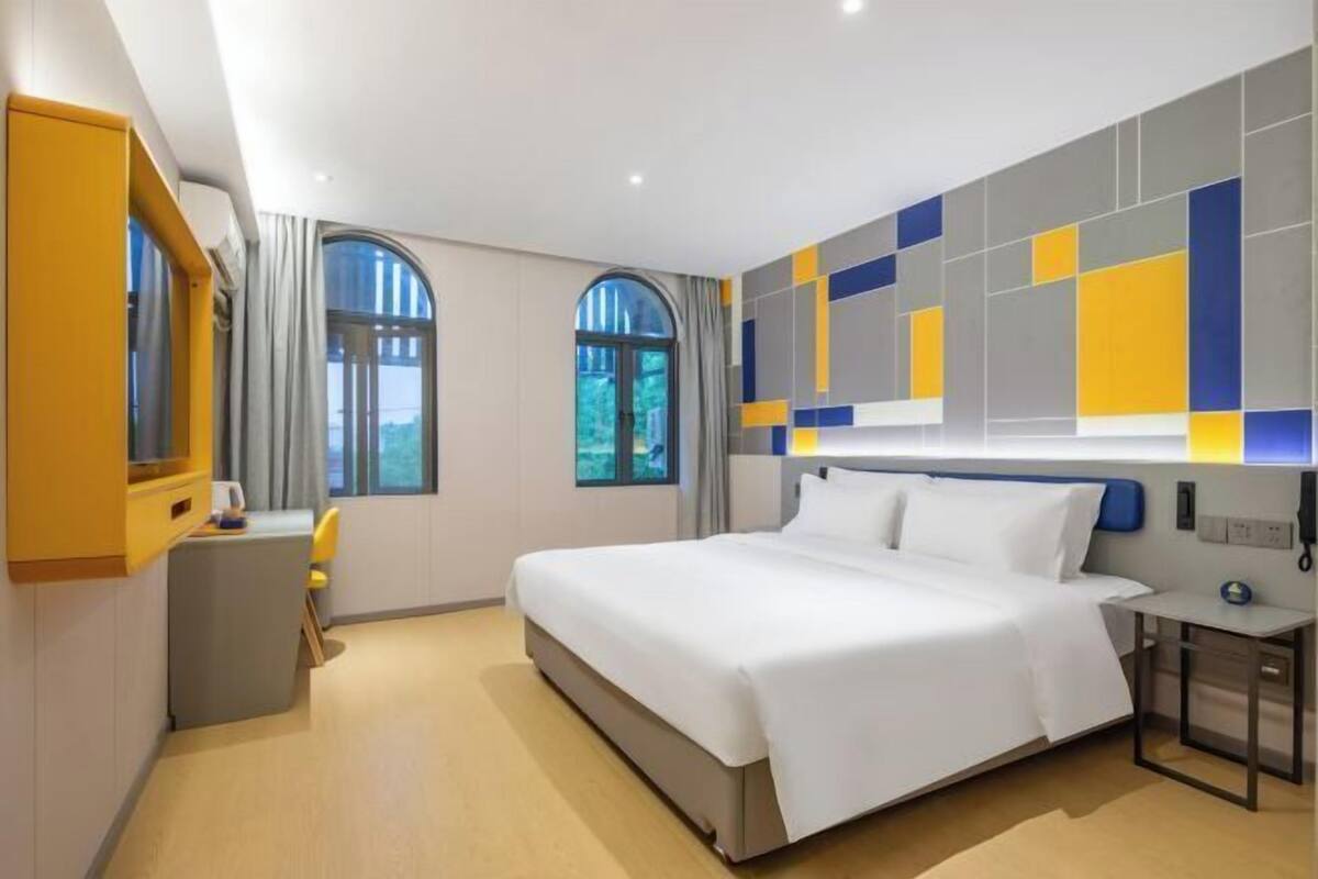 A modern hotel room with a large bed, two windows, a desk, and a wall with geometric patterns.