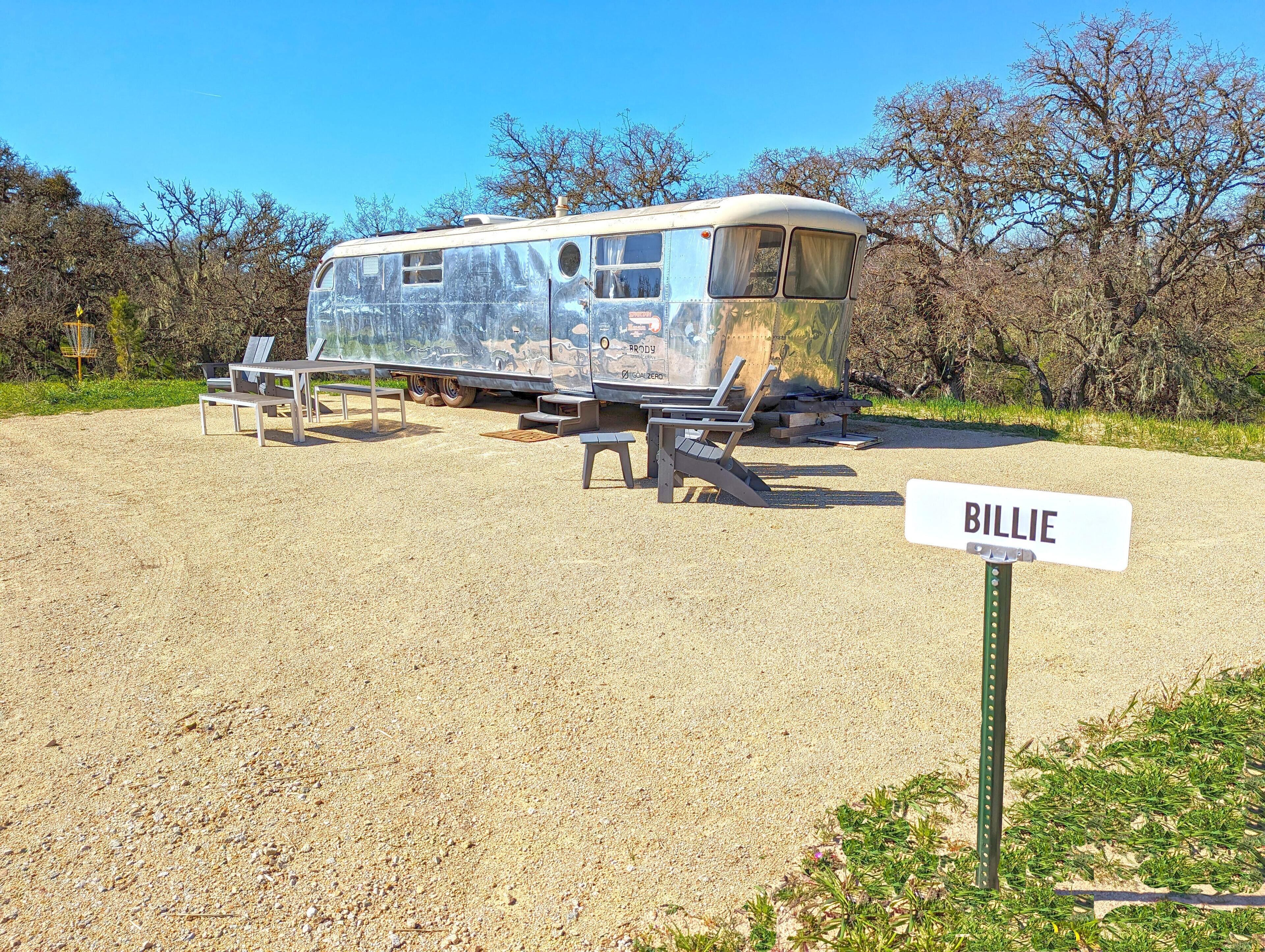 A vintage camper with a "Billie" sign, surrounded by outdoor seating and a gravel area.