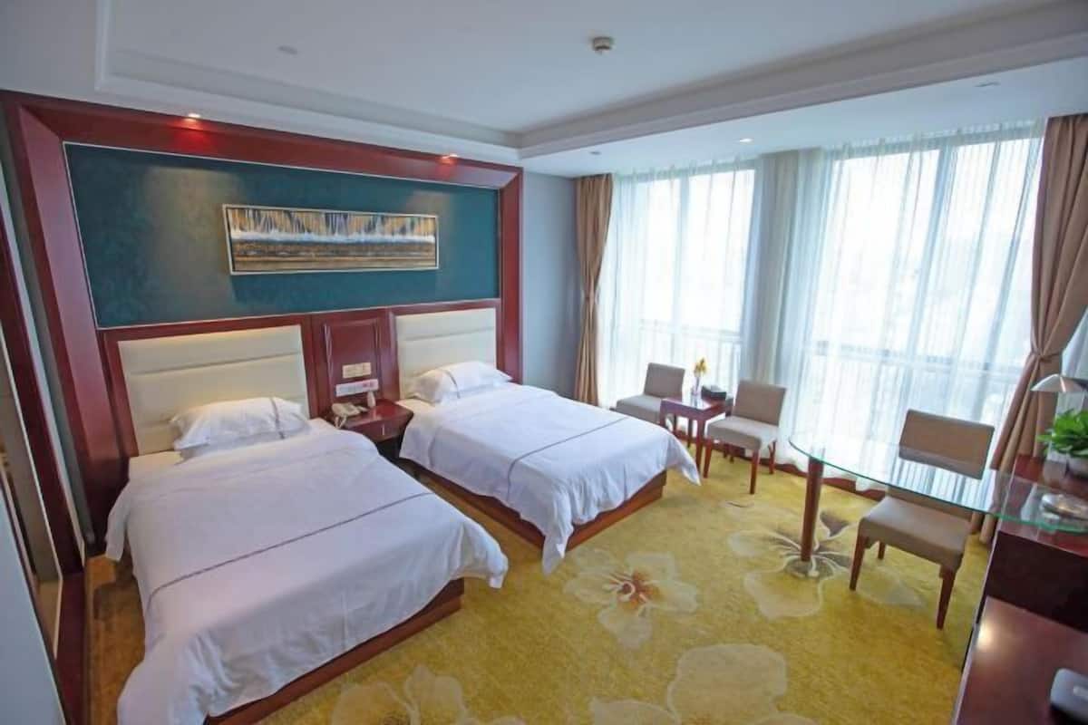 A hotel room with two beds, a desk, a chair, and a large window with curtains.