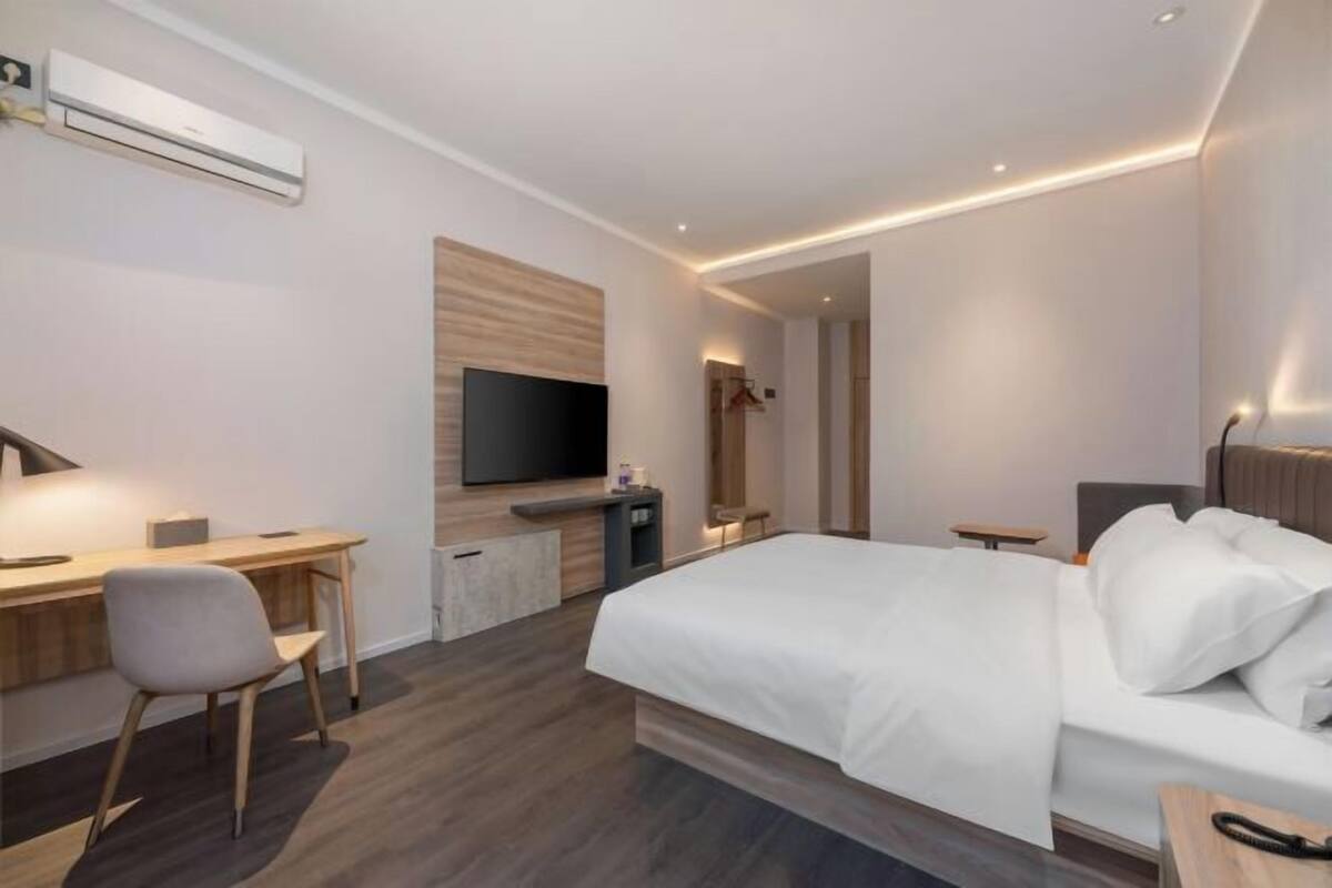 A modern hotel room with a bed, desk, chair, TV, and an air conditioning unit.