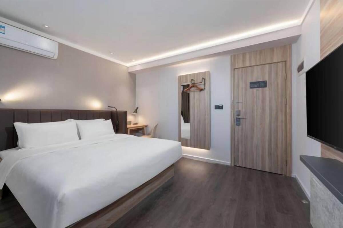 A modern hotel room with a large bed, a desk with a lamp, a mirror, and a TV mounted on the wall.
