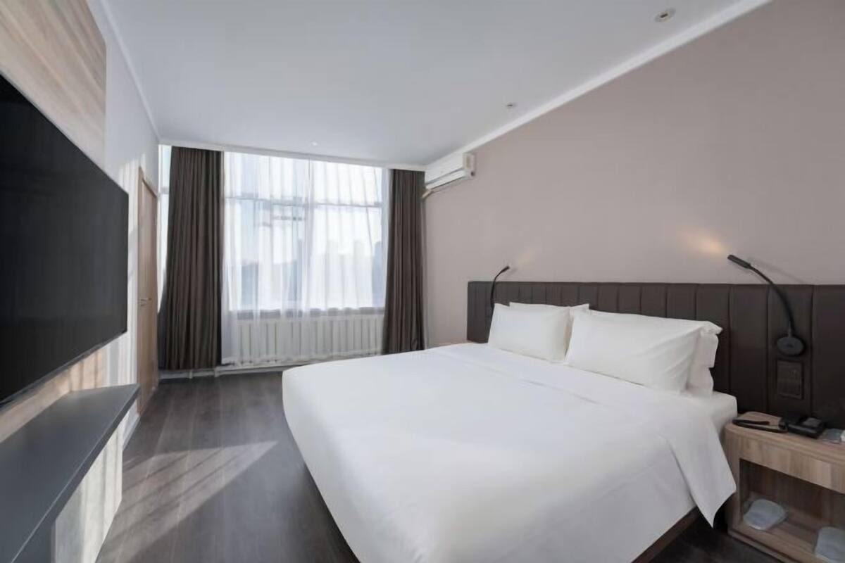 A modern hotel room with a large bed, a flat-screen TV, a bedside table with a lamp, and a view of the city through large windows.