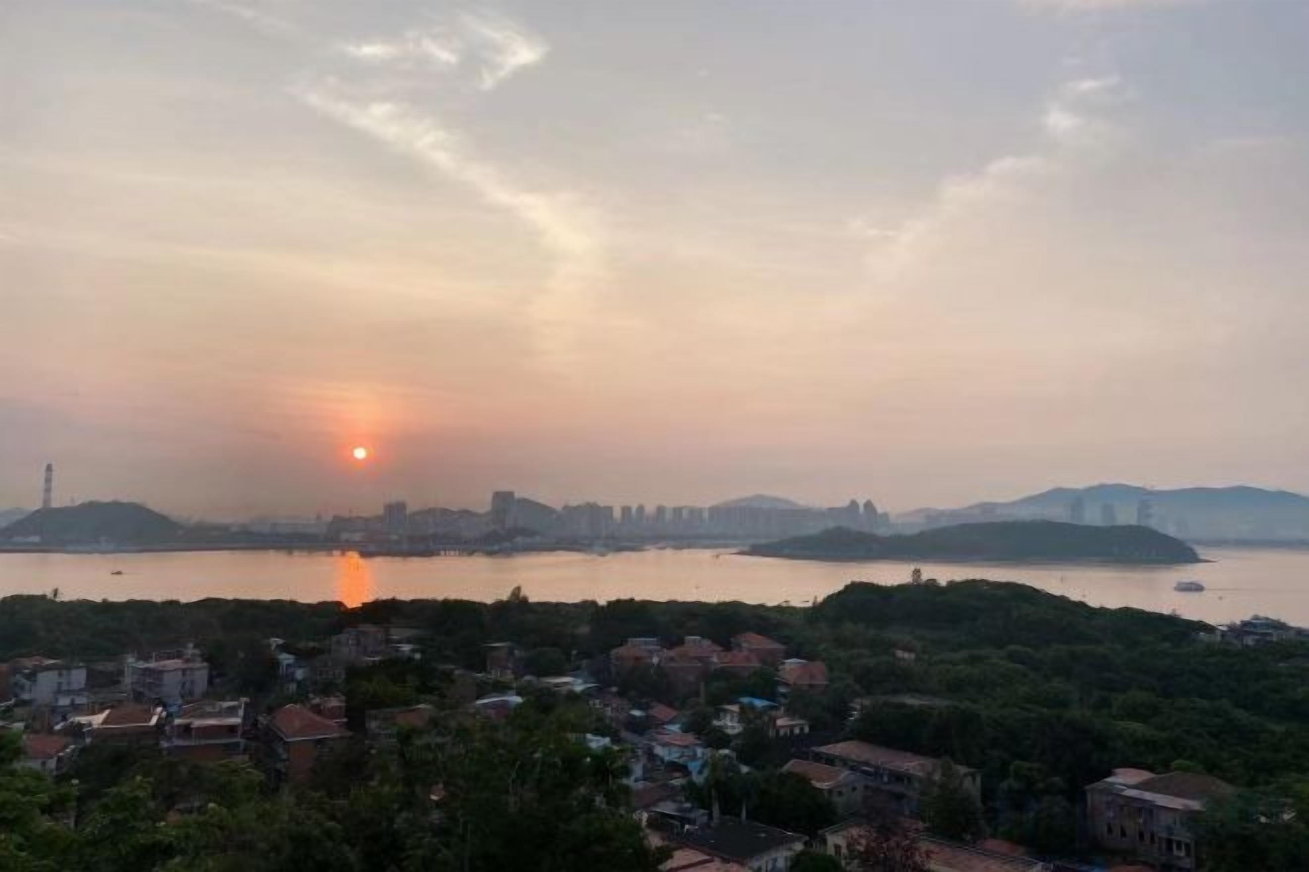 Un paisaje urbano con un atardecer sobre una masa de agua, edificios en primer plano y colinas al fondo.