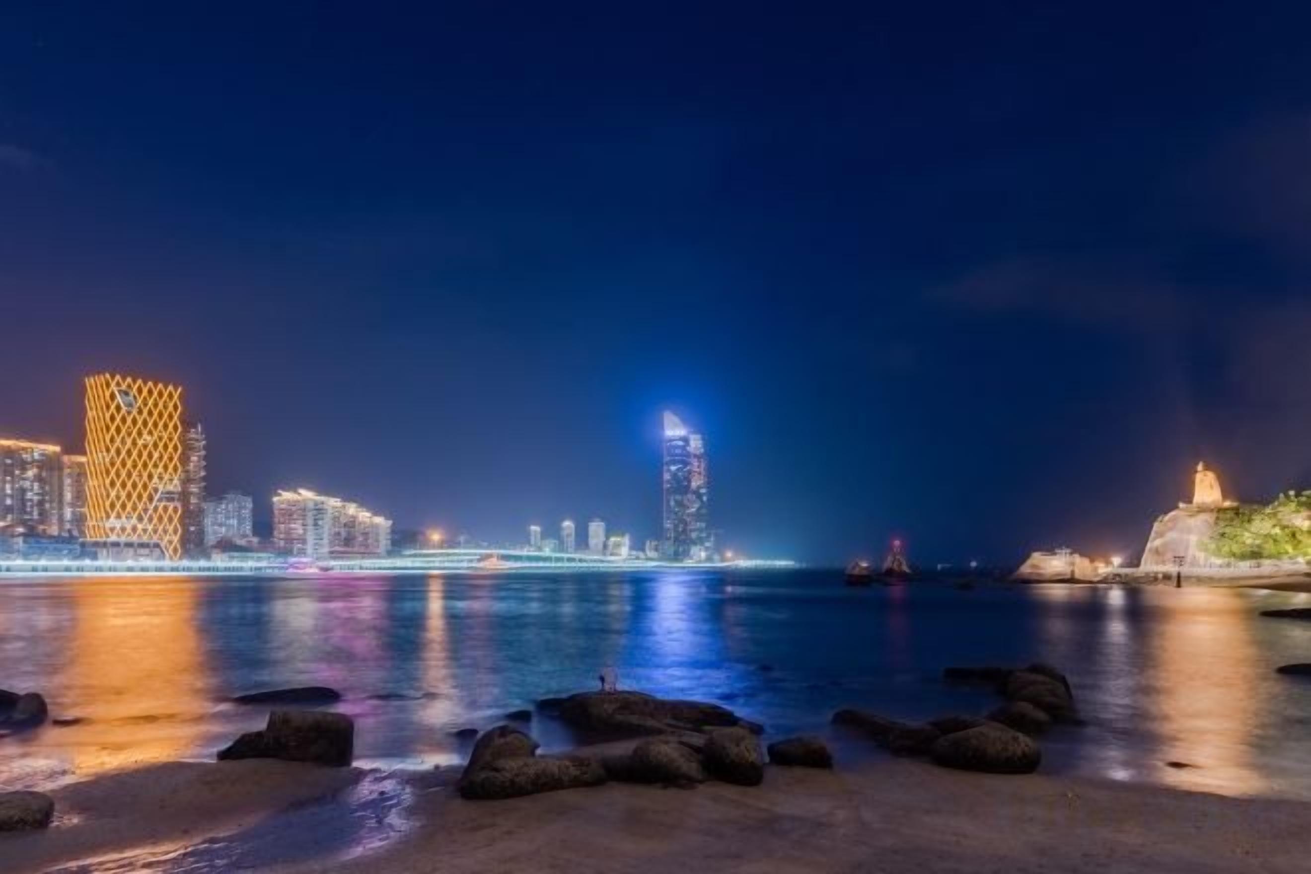 A nighttime cityscape with illuminated buildings and a calm waterfront.
