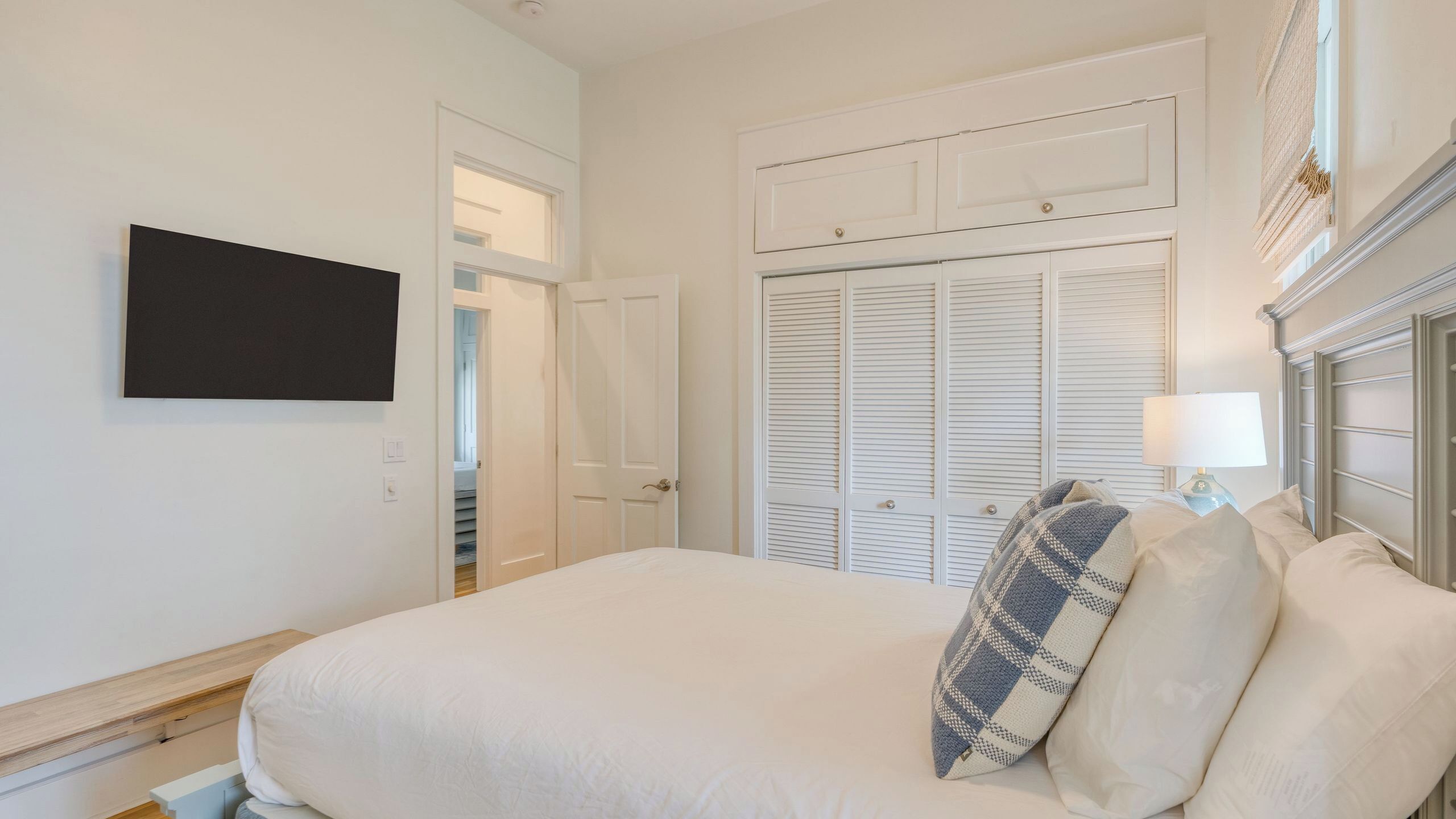 A bedroom with a bed, a television mounted on the wall, a closet with louvered doors, and a bedside lamp.
