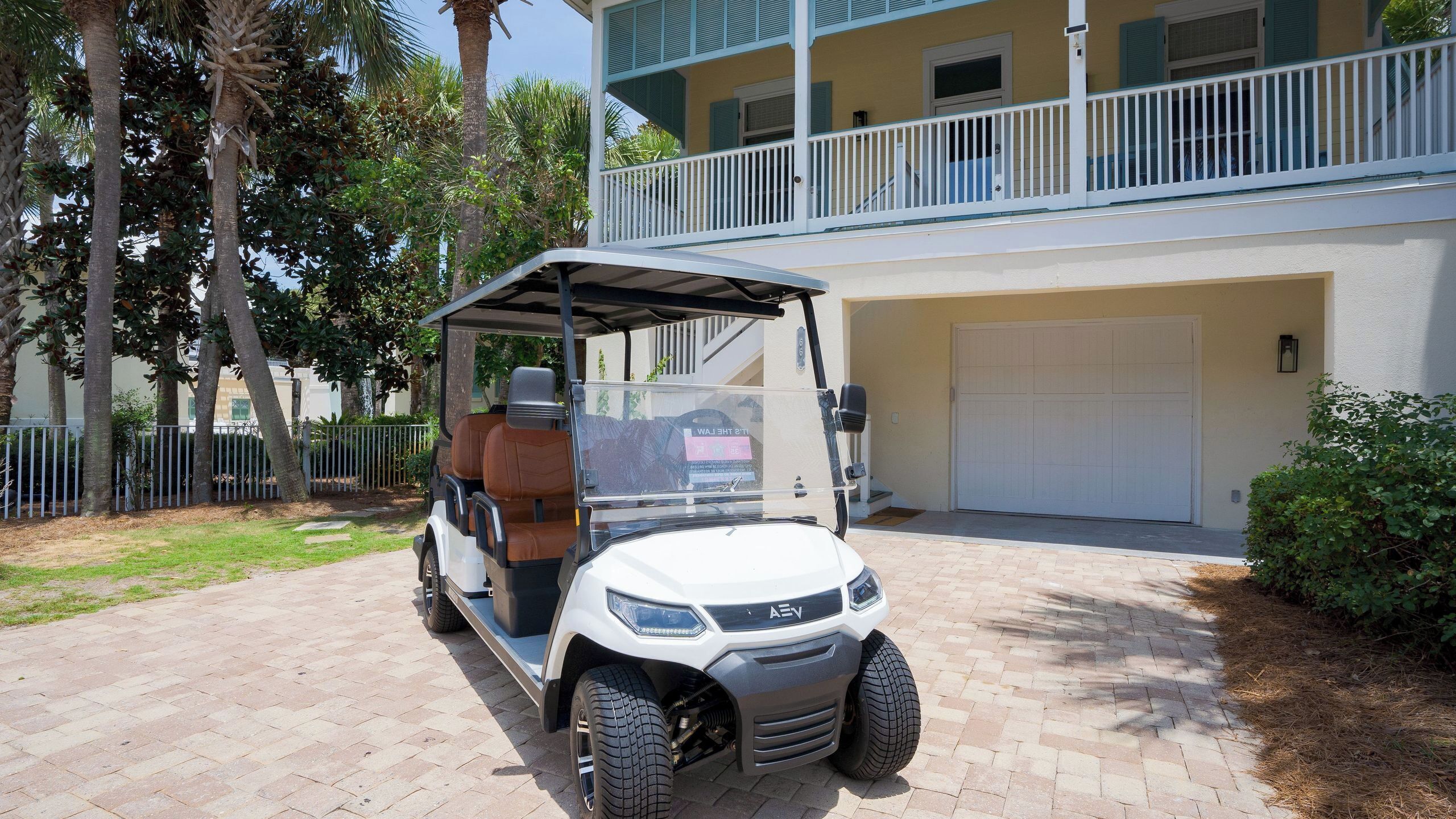 A golf cart parked in front of a building with a garage, a balcony, and a fenced area with palm trees.